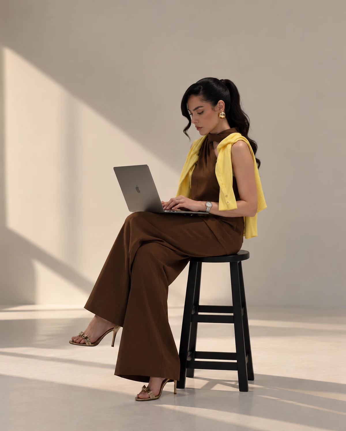 Sabrina Reis sentada em um banco com laptop, em estúdio iluminado pelo sol.